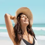 A smiling woman with glowing skin at the beach wearing a sun hat