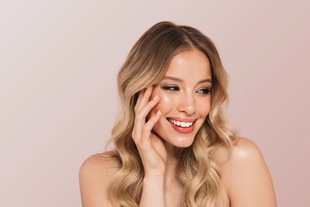 A smiling woman with clear skin, touching her face with her hands against a pink background