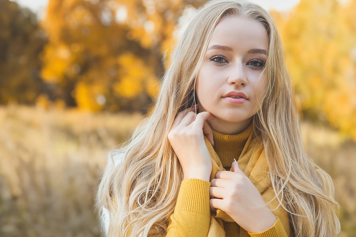 A woman in a yellow sweater with fall trees and yellow, orange, and red foliage out of focus on the background