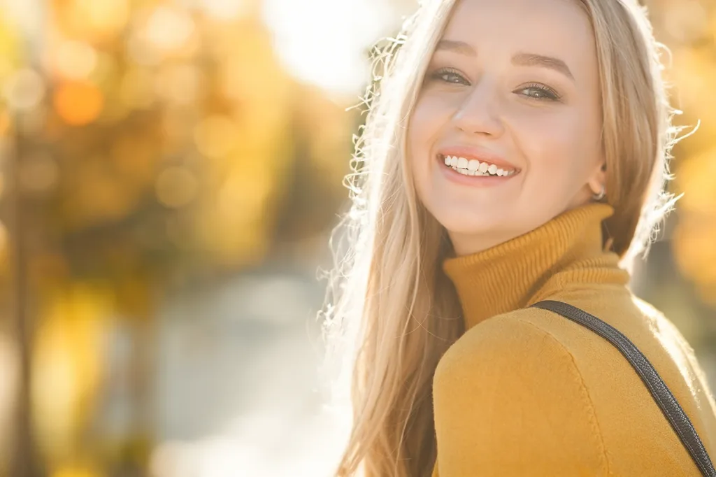 A smiling woman in a yellow sweater with fall trees and yellow, orange, and red foliage out of focus on the background