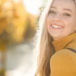 A smiling woman in a yellow sweater with fall trees and yellow, orange, and red foliage out of focus on the background