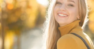 A smiling woman in a yellow sweater with fall trees and yellow, orange, and red foliage out of focus on the background