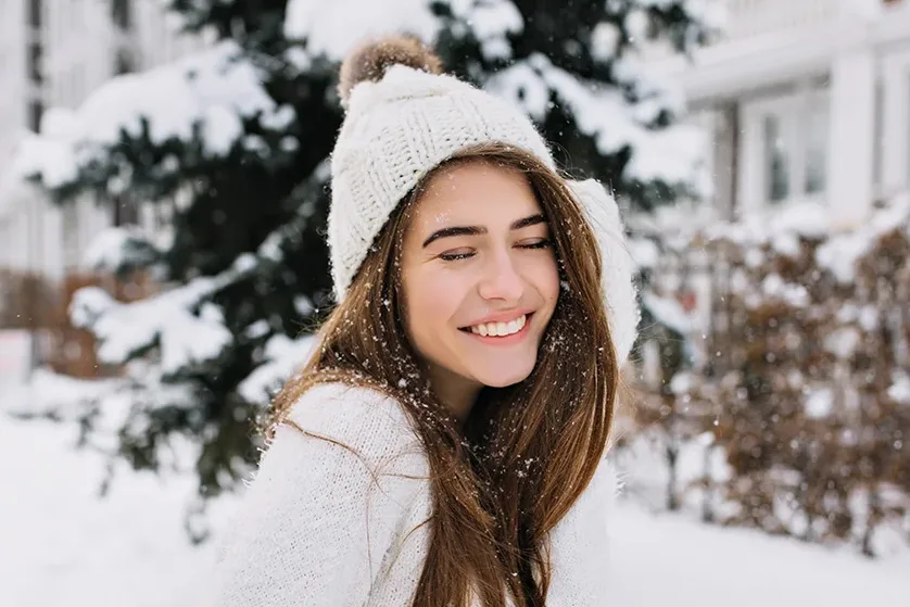 A young smiling woman stands outside in the snow, wearing a knit sweater and hat