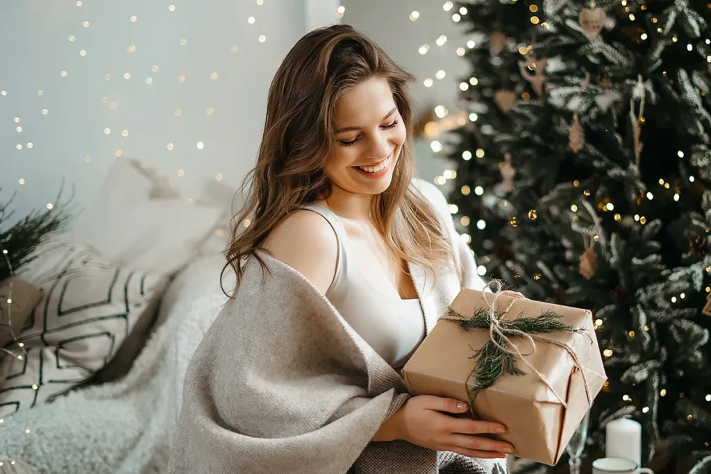 A smiling woman holds a gift, surrounded by holiday lights and seasonal winter decor