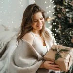 A smiling woman holds a gift, surrounded by holiday lights and seasonal winter decor