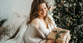 A smiling woman holds a gift, surrounded by holiday lights and seasonal winter decor